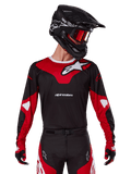 Alpinestars 2025 Racer Veil Jersey, koszulka motocrossowa, czarno-jaskrawoczerwona, noszona z dopasowanymi spodniami, czarnym kaskiem i czerwonymi goglami, wyraziste białe logo i nowoczesny wyścigowy design, profesjonalna odzież do motocrossu terenowego