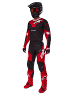 Zawodnik motocrossowy w kompletnym stroju wyścigowym Alpinestars 2025 Racer Veil w kolorze czarnym i jasnoczerwonym, pasujące spodnie, czerwone buty, czarny kask z czerwonymi goglami oraz czerwono-czarne rękawice, profesjonalny zestaw do wyścigów terenowych