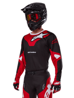 Motocyklista motocrossowy w zestawie Alpinestars 2025 Racer Veil, czerwono-czarna koszulka z białym logo Alpinestars, pasujące spodnie i rękawice, czarny kask z czerwonymi goglami, techniczna odzież do wyścigów off-road