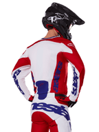 Alpinestars 2025 Racer Veil Jersey and Pants, zestaw odzieży motocrossowej, jaskrawoczerwony, biały i niebieski, widok z tyłu motocyklisty w pasującym zestawie i czarnym kasku motocrossowym, wyraźne niebieskie logo Alpinestars, techniczna odzież wyścigowa off-road