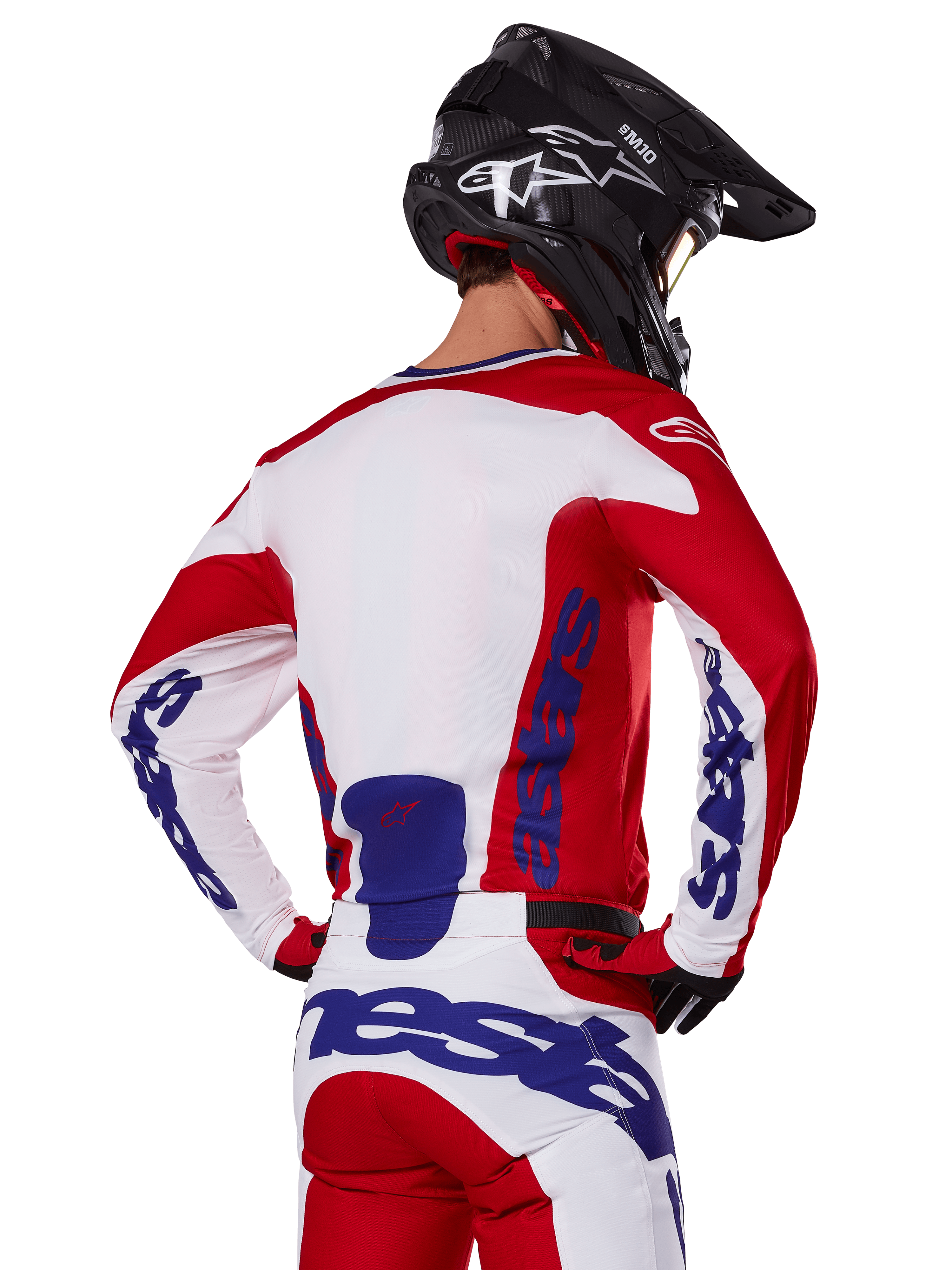 Alpinestars 2025 Racer Veil Jersey and Pants, zestaw odzieży motocrossowej, jaskrawoczerwony, biały i niebieski, widok z tyłu motocyklisty w pasującym zestawie i czarnym kasku motocrossowym, wyraźne niebieskie logo Alpinestars, techniczna odzież wyścigowa off-road