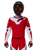 Alpinestars 2025 Racer Veil Jersey, koszulka motocrossowa z długim rękawem, czerwona, biała i niebieska, noszona z czarnym kaskiem i pomarańczowymi goglami, pasujące czerwone rękawice i spodnie, nowoczesny design do jazdy terenowej