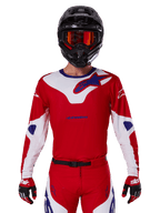 Alpinestars 2025 Racer Veil Jersey, koszulka motocrossowa z długim rękawem, czerwona, biała i niebieska, noszona z czarnym kaskiem i pomarańczowymi goglami, pasujące czerwone rękawice i spodnie, nowoczesny design do jazdy terenowej