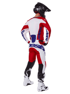 Motocyklista motocrossowy widoczny od tyłu, ubrany w koszulkę i spodnie Alpinestars 2025 Racer Veil w kolorze czerwonym i białym, niebieski branding, czarny kask i białe buty, kompletny zestaw odzieży do wyścigów terenowych