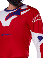 Alpinestars 2025 Racer Veil Jersey, koszulka motocrossowa, jaskrawoczerwona i biała, zbliżenie na klatkę piersiową i górną część tułowia, niebieskie logo Alpinestars na białym panelu kołnierza, czerwone rękawy z białymi akcentami, noszona z czarnym kaskiem, techniczna odzież do jazdy terenowej