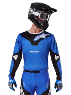 Koszulka 2025 Racer Veil, koszulka motocrossowa, niebieska i czarna, noszona z dopasowanym sprzętem motocrossowym, w tym kaskiem i rękawicami, sportowy design z białym brandingiem Alpinestars