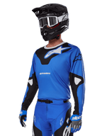 Zestaw Alpinestars 2025 Racer Veil, niebieska koszulka motocrossowa z czarno-białymi grafikami, noszona z pasującymi spodniami, czarnym kaskiem, białymi goglami i niebieskimi rękawicami, techniczna odzież do wyścigów terenowych