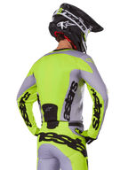 Zawodnik motocrossowy od tyłu w koszulce Alpinestars 2025 Racer Veil w kolorze szarym i neonowym żółtym, z wyraźnym logo „astars” na plecach i rękawach, w zestawie z dopasowanymi spodniami i czarnym kaskiem, kompletny strój wyścigowy