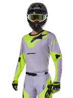 Zawodnik motocrossowy w zestawie Alpinestars 2025 Racer Veil w kolorze szarym i neonowym zielonym, jasnoszara koszulka i spodnie z wyrazistymi neonowymi żółto-zielonymi panelami i czarnym logo Alpinestars, dopasowane rękawice i czarny kask z neonowymi zielonymi goglami, profesjonalna odzież wyścigowa off-road.