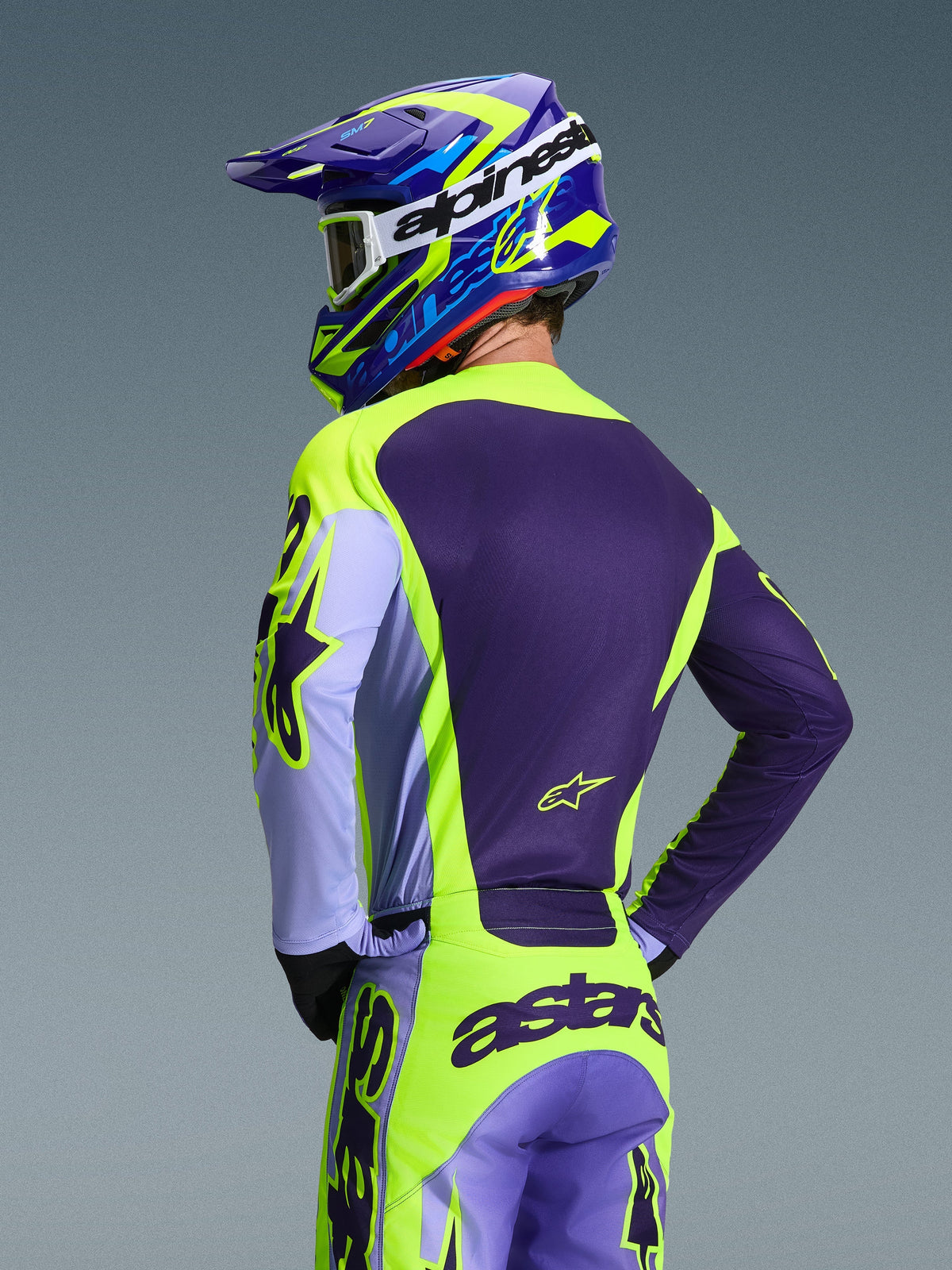 Kierowca motocrossowy widziany od tyłu w koszulce Alpinestars 2026 Racer Portl i pasujących spodniach, fioletowo-żółta fluorescencyjna kolorystyka z lawendowymi akcentami, dopasowany niebiesko-żółty kask, odzież o sportowym kroju race-fit zaprojektowana do jazdy off-road