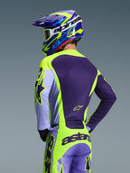 Kierowca motocrossowy widziany od tyłu w koszulce Alpinestars 2026 Racer Portl i pasujących spodniach, fioletowo-żółta fluorescencyjna kolorystyka z lawendowymi akcentami, dopasowany niebiesko-żółty kask, odzież o sportowym kroju race-fit zaprojektowana do jazdy off-road