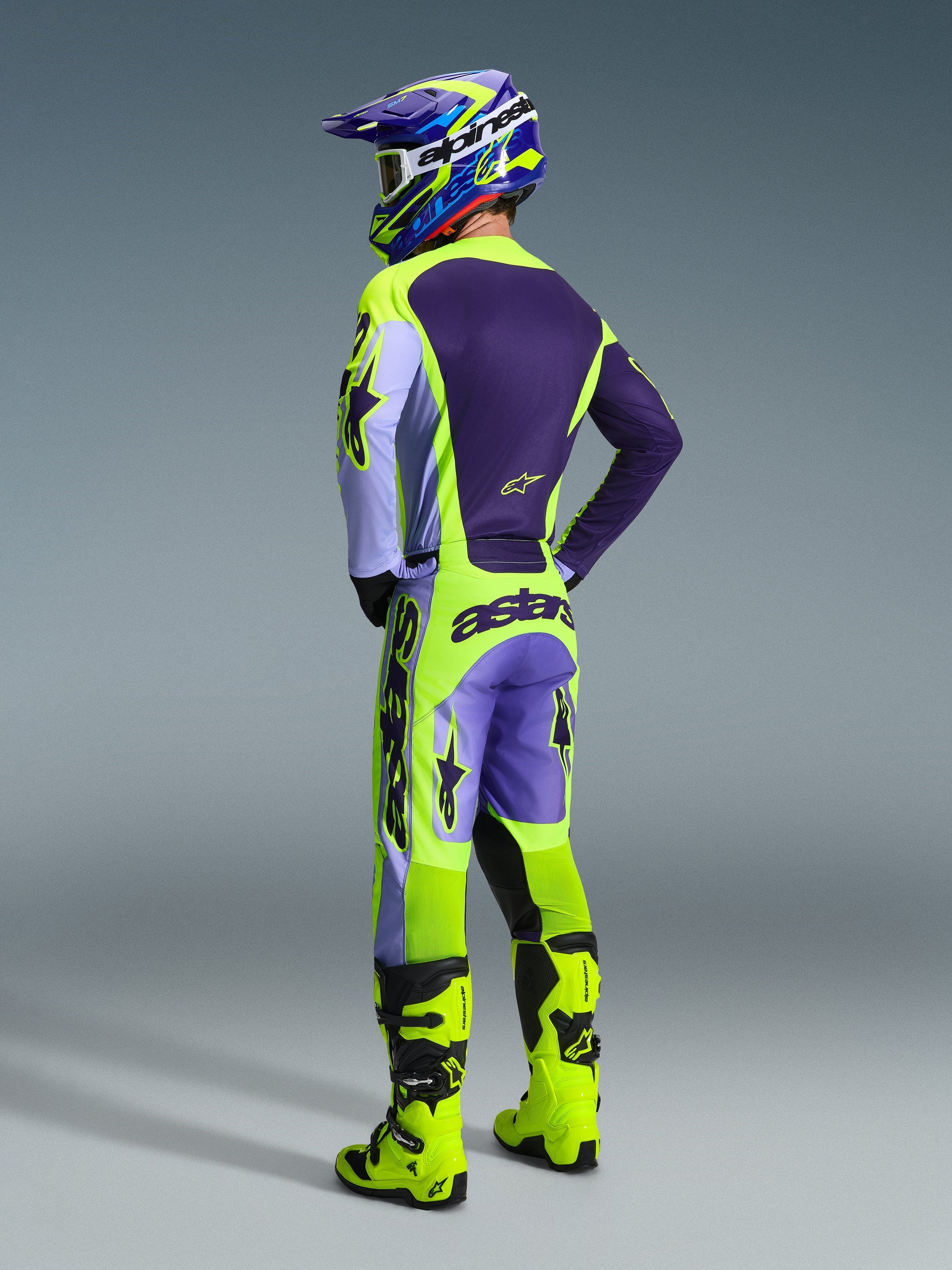 Zawodnik motocrossowy w koszulce i spodniach Alpinestars 2026 Racer Portl, fioletowy i neonowy żółty zestaw odzieży, widok od tyłu, dopasowany kask i neonowożółte buty, profesjonalny strój do wyścigów terenowych