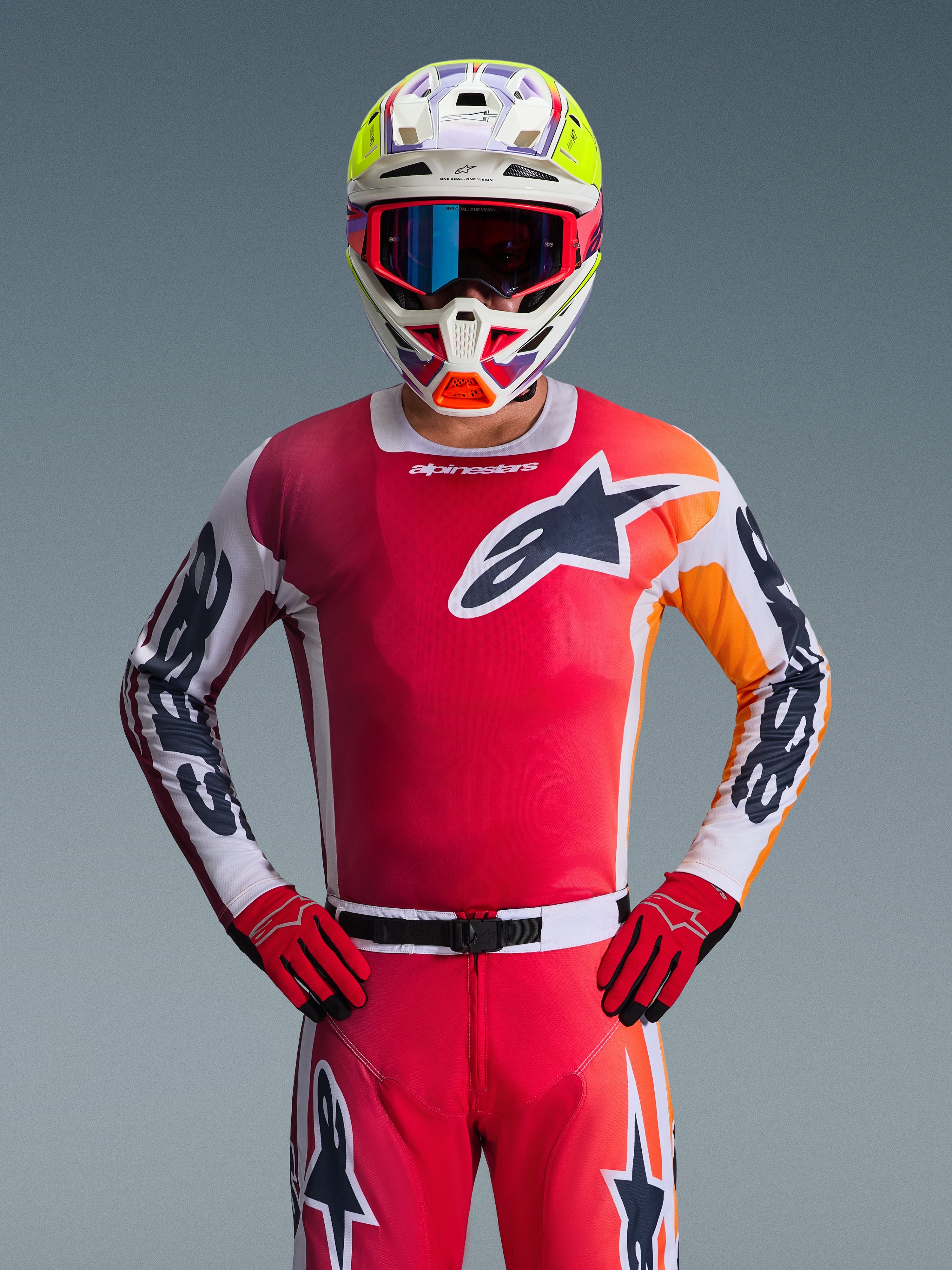 Zestaw Alpinestars 2026 Racer Portl, zawodnik motocrossowy w jaskrawoczerwonej koszulce z pomarańczowymi i białymi akcentami, pasujące czerwone spodnie i rękawice, wielokolorowy kask z neonowożółtymi elementami i niebieskimi goglami, duże logo z gwiazdą na piersi, profesjonalny strój do wyścigów off-road