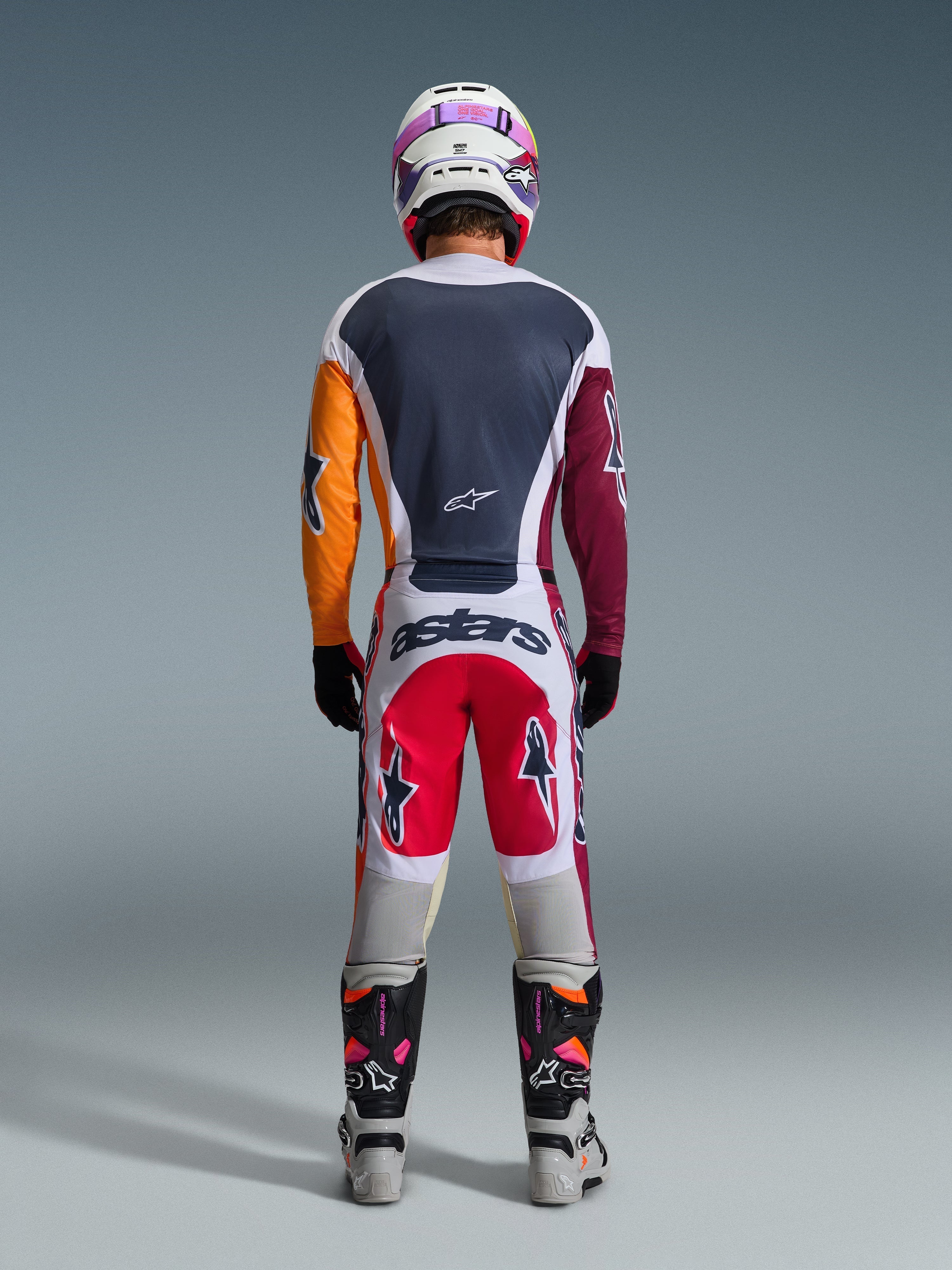 Motocrossista Alpinestars widoczny od tyłu w kompletnym stroju wyścigowym, koszulka 2026 Racer Portl w kolorach czerwonym, jasnoszarym i ciemnozielonym z asymetrycznymi pomarańczowymi i bordowymi rękawami, dopasowane biało-czerwone spodnie, biały kask z fioletowymi goglami, biało-czarne buty, skoordynowany zestaw do jazdy off-road