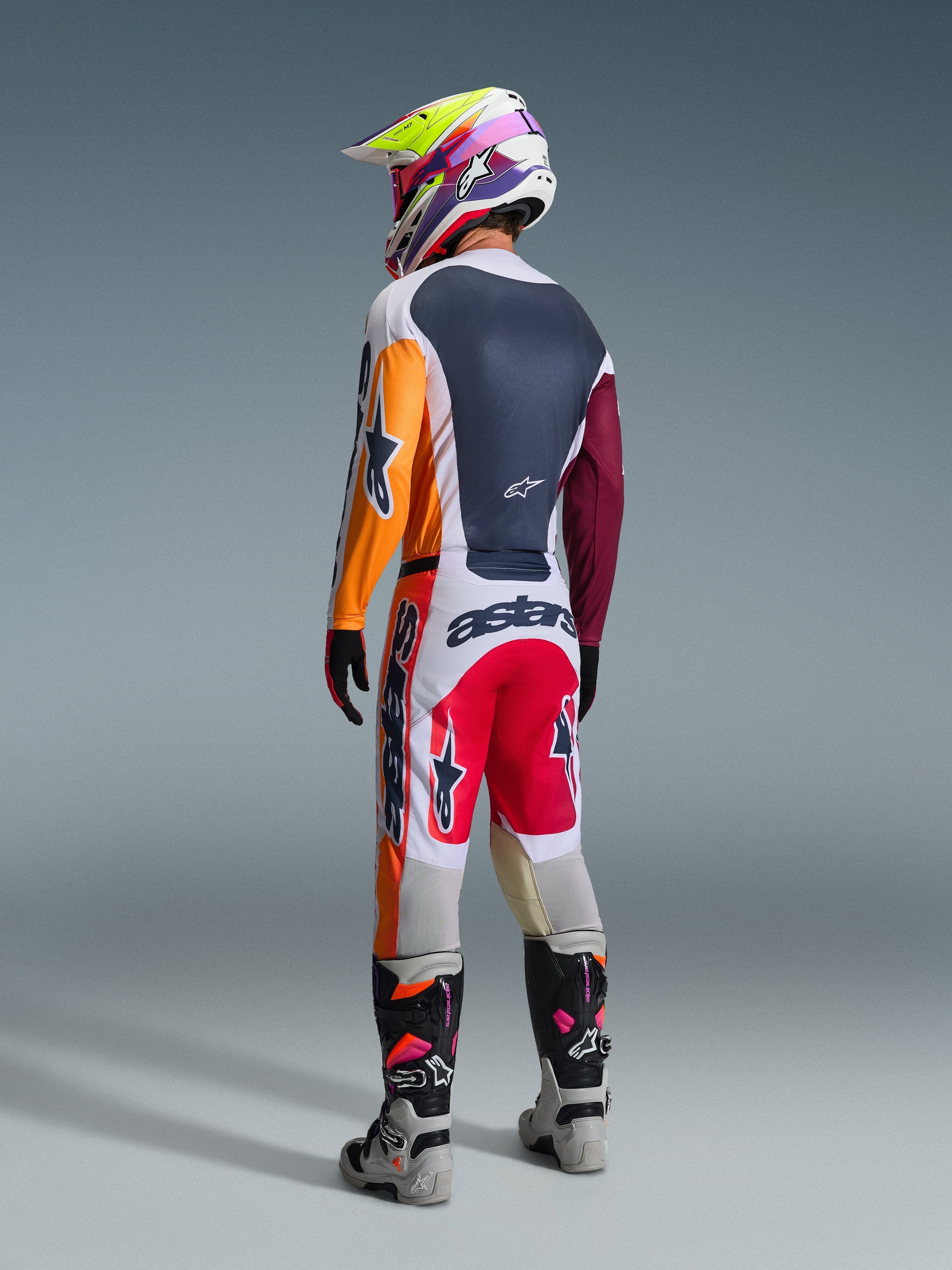 Zawodnik motocrossowy widziany z tyłu w zestawie Alpinestars 2026 Racer Portl, koszulka z pomarańczowymi i bordowymi rękawami, białe spodnie z czerwonymi panelami i wyraźnym logo „astars”, wielokolorowy kask, szare buty, profesjonalny strój wyścigowy off-road