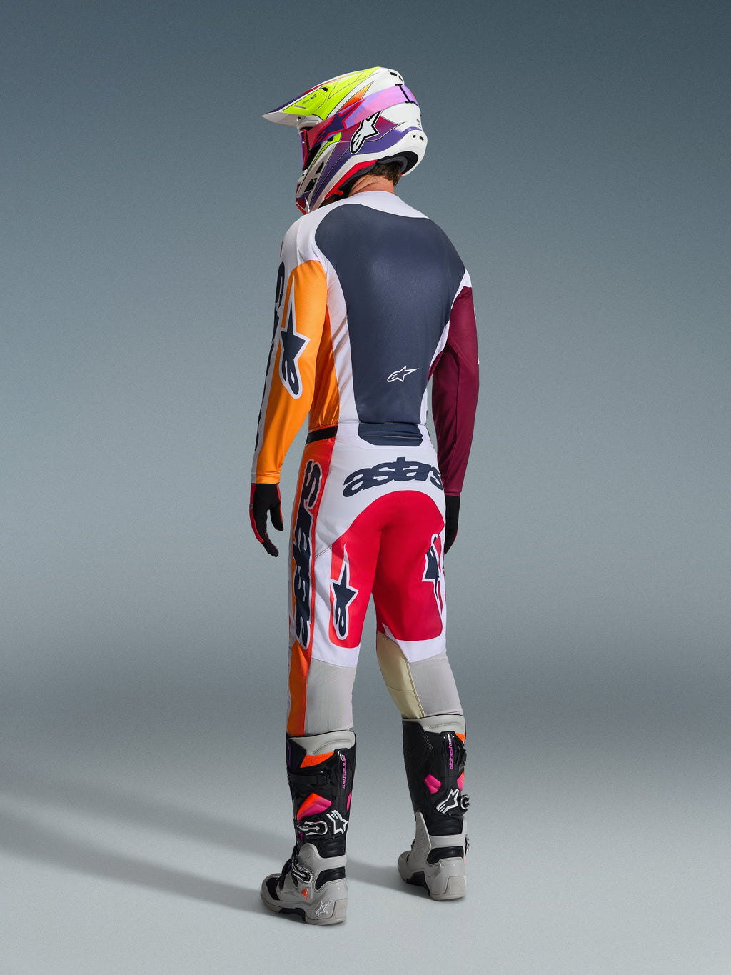 Zawodnik motocrossowy w pełnym stroju wyścigowym widziany od tyłu, koszulka Alpinestars 2026 Racer Portl w kolorze czerwono-jasnoszarym i ciemnozielonym z pomarańczowymi i bordowymi panelami, pasujące biało-czerwone spodnie, szaro-czarne buty z różowymi detalami, wielokolorowy kask, spójny zestaw do wyścigów off-road