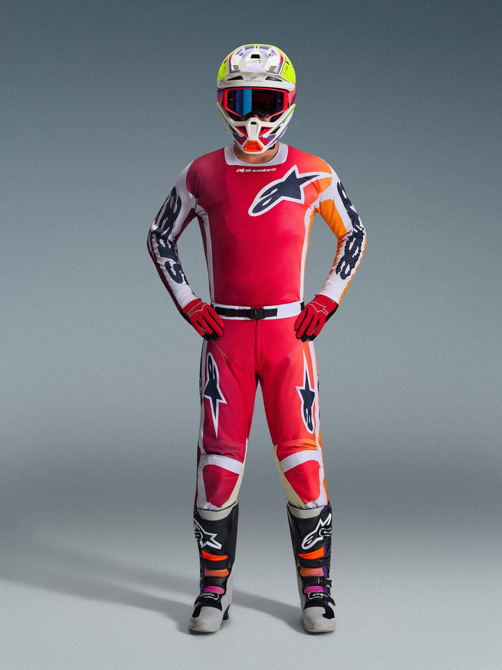 Motocyklista motocrossowy w koszulce Alpinestars 2026 Racer Portl w kolorze czerwonym z białymi i pomarańczowymi akcentami graficznymi, dopasowane spodnie motocrossowe, czerwone rękawice, kolorowy kask wyścigowy z goglami i czarno-białe buty techniczne, kompletny zestaw odzieży off-road