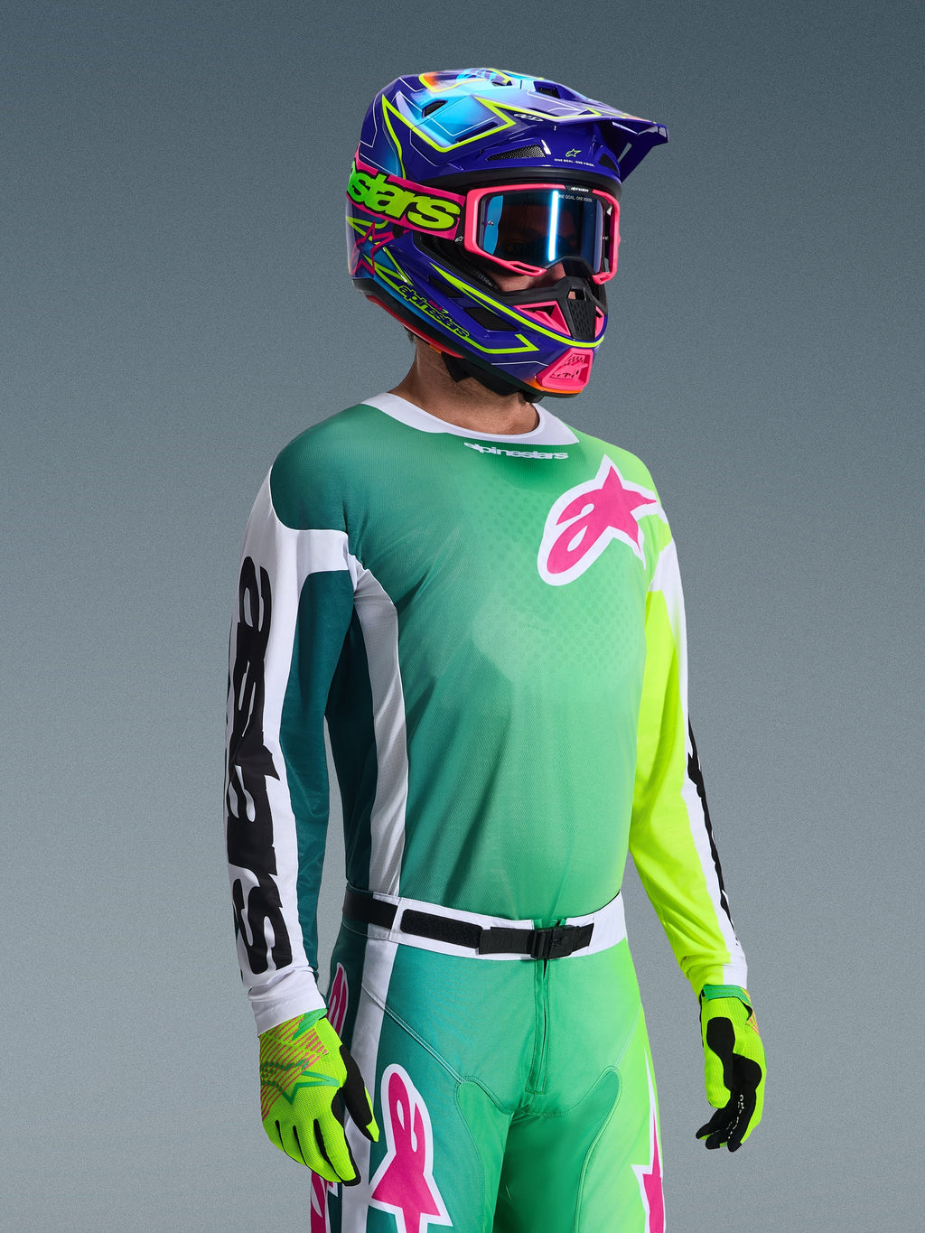 Alpinestars 2026 Racer Portl Jersey, koszulka motocrossowa, zielono-biało-czarna, noszona przez zawodnika z dopasowanymi zielonymi spodniami, fluorescencyjnymi rękawicami oraz intensywnie niebiesko-fioletowym kaskiem z różowymi goglami, ozdobiona różowym logo Astars na klatce piersiowej