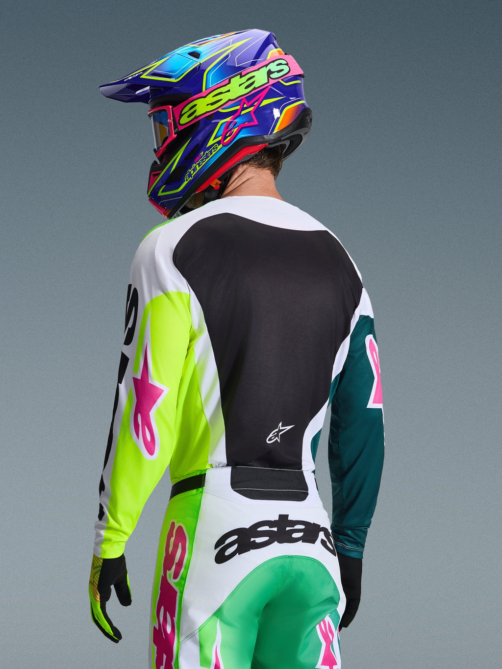 Zawodnik motocrossowy widziany od tyłu w koszulce Alpinestars 2026 Racer Portl w kolorze zielono-biało-czarnym, z różowymi logotypami gwiazdy, w zestawie z dopasowanymi spodniami i kolorowym niebiesko-fioletowym kaskiem, profesjonalny strój wyścigowy off-road.