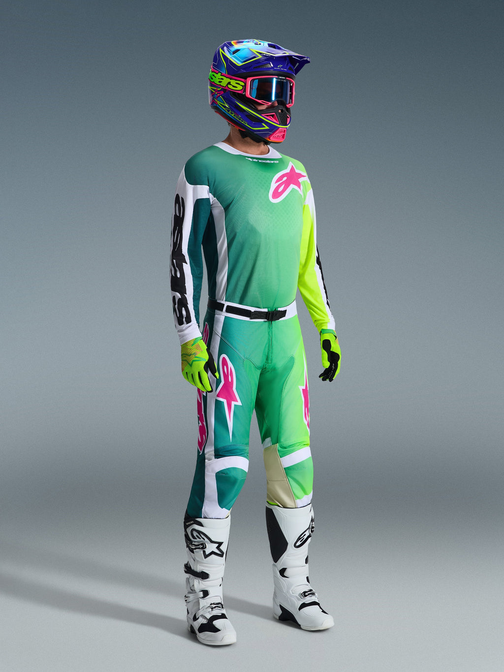 Zawodnik motocrossowy Alpinestars w pełnym stroju wyścigowym, zielona koszulka Racer Portl 2026 z biało-czarnymi rękawami i różowym logo, pasujące zielone spodnie z różowymi akcentami, wielokolorowy kask z goglami, biało-czarne buty i zielone rękawice, skoordynowany zestaw do jazdy off-road