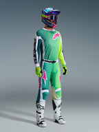 Zawodnik motocrossowy Alpinestars w pełnym stroju wyścigowym, zielona koszulka Racer Portl 2026 z biało-czarnymi rękawami i różowym logo, pasujące zielone spodnie z różowymi akcentami, wielokolorowy kask z goglami, biało-czarne buty i zielone rękawice, skoordynowany zestaw do jazdy off-road