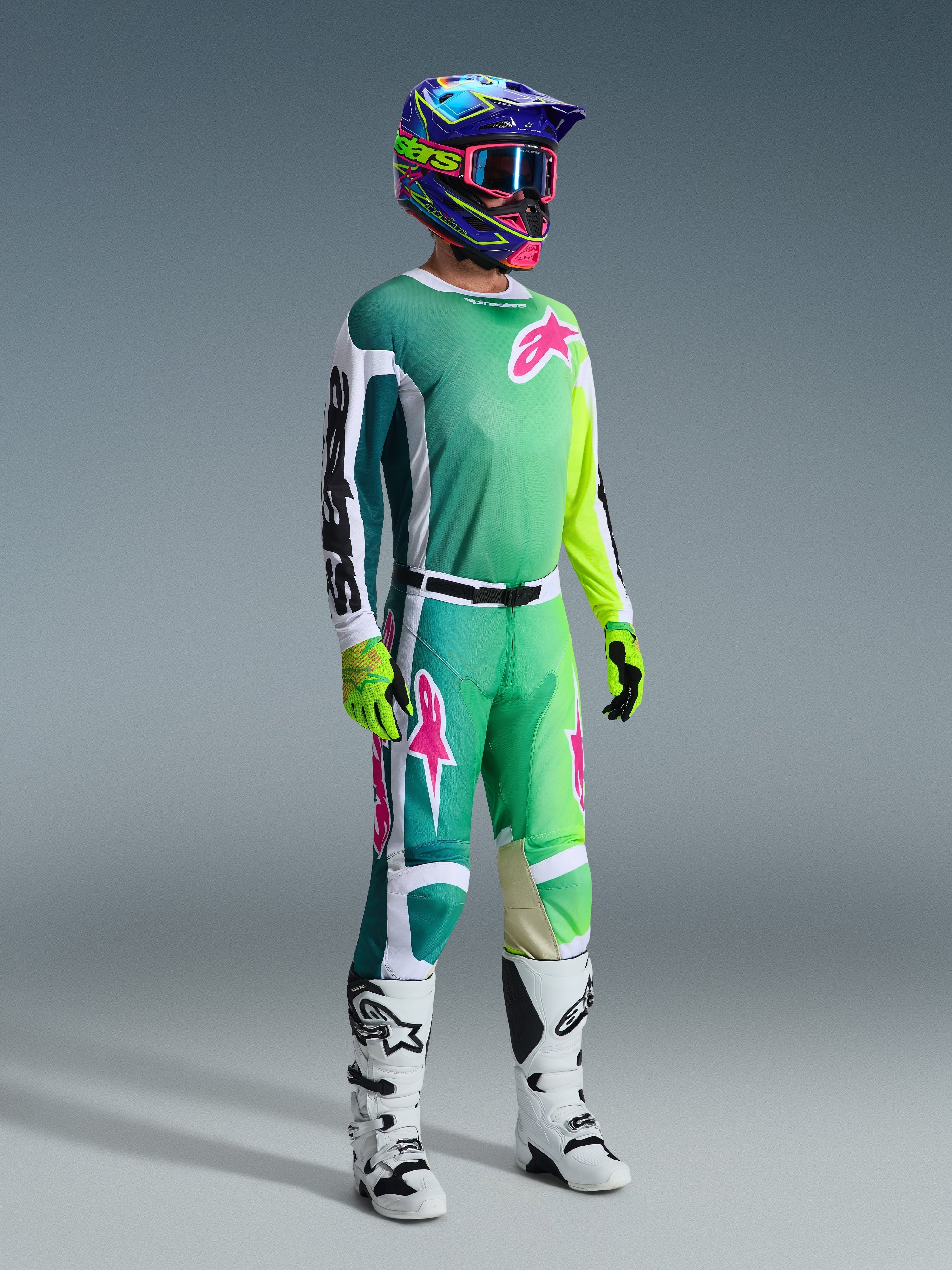 Zawodnik motocrossowy Alpinestars w pełnym stroju wyścigowym, zielona koszulka Racer Portl 2026 z biało-czarnymi rękawami i różowym logo, pasujące zielone spodnie z różowymi akcentami, wielokolorowy kask z goglami, biało-czarne buty i zielone rękawice, skoordynowany zestaw do jazdy off-road
