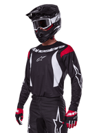 Motocyklista motocrossowy ubrany w zestaw Alpinestars 2025 Fluid Haul, czarna koszulka z czerwonymi panelami na ramionach i białą grafiką, pasujące czarne spodnie z białym logo gwiazdy, czerwono-czarne rękawice, czarny kask z białymi akcentami i gogle, dopasowana odzież motocrossowa.