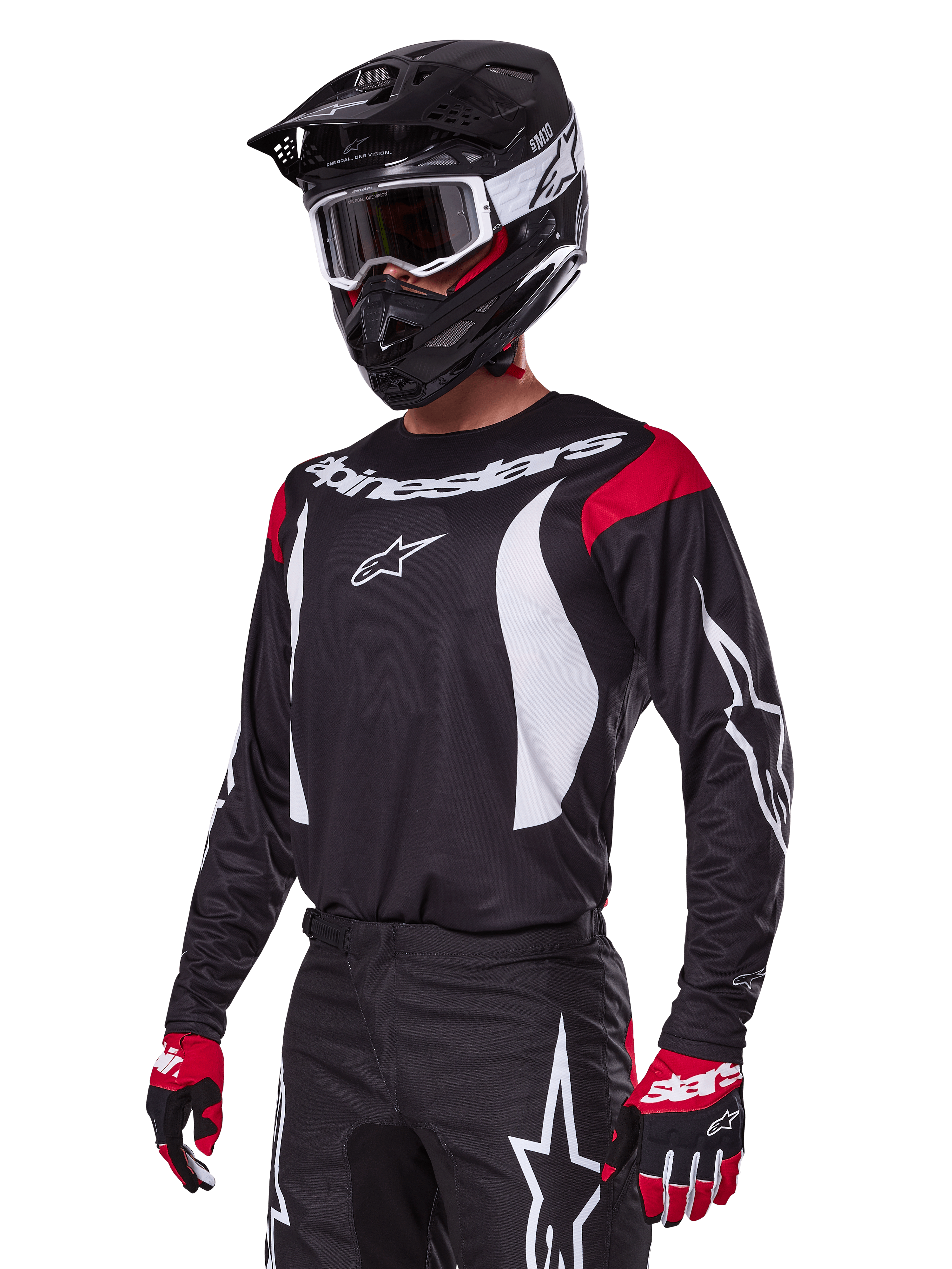 Motocyklista motocrossowy ubrany w zestaw Alpinestars 2025 Fluid Haul, czarna koszulka z czerwonymi panelami na ramionach i białą grafiką, pasujące czarne spodnie z białym logo gwiazdy, czerwono-czarne rękawice, czarny kask z białymi akcentami i gogle, dopasowana odzież motocrossowa.