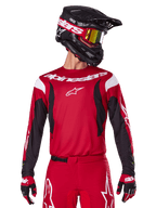 Alpinestars 2025 Fluid Haul Jersey, koszulka motocrossowa, jaskrawoczerwona i czarna, noszona jako część kompletnego zestawu z pasującymi spodniami i rękawicami, czarny kask z goglami z żółtymi szybkami, białe logo i branding, wysokiej klasy odzież do wyścigów off-road