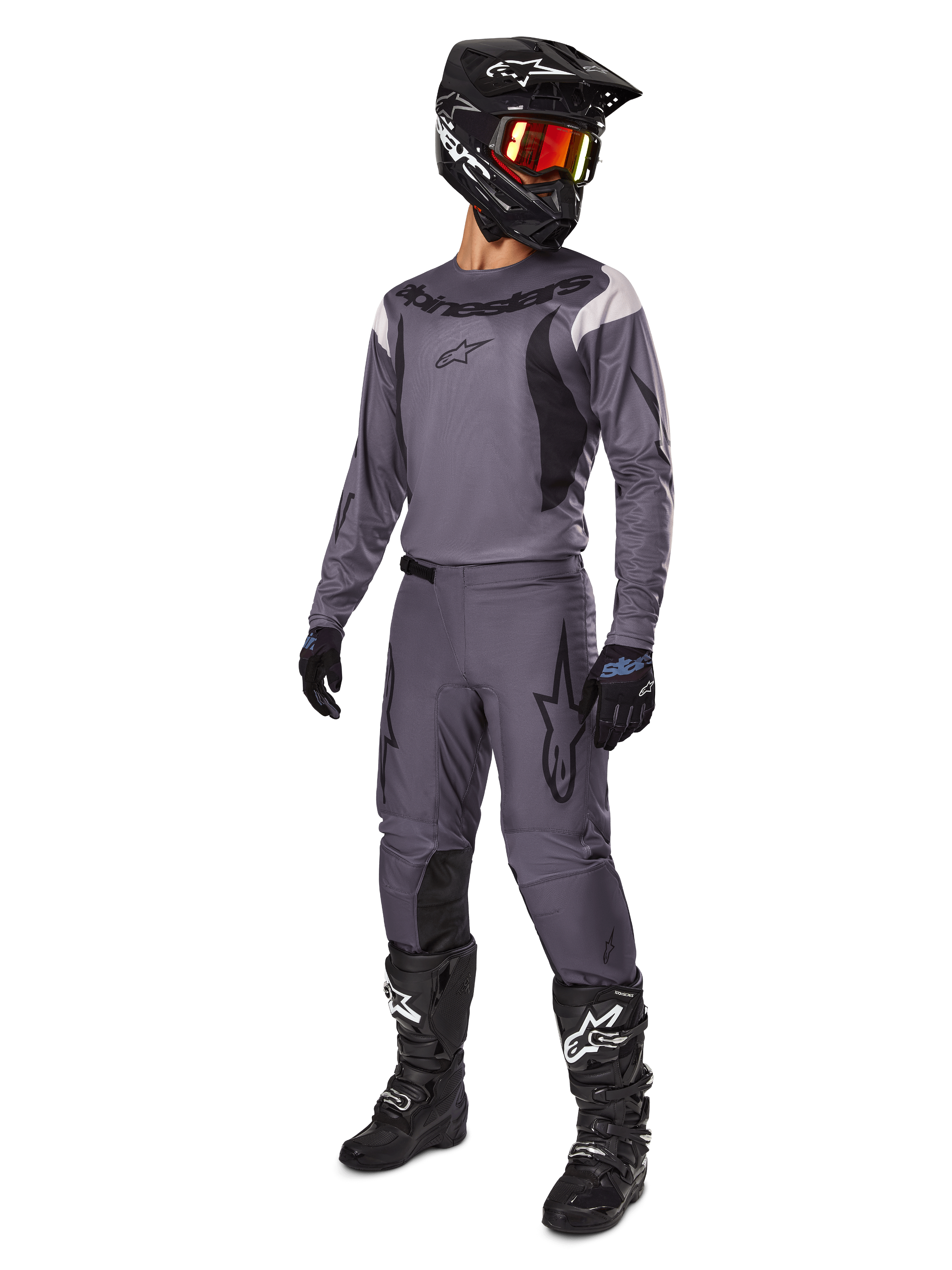 Motocyklista motocrossowy w szarym zestawie Alpinestars 2025 Fluid Haul, szara koszulka i spodnie z czarnymi akcentami, czarny kask z czerwonawymi goglami, czarne rękawice i czarne buty z białym logo, kompletny strój do wyścigów terenowych