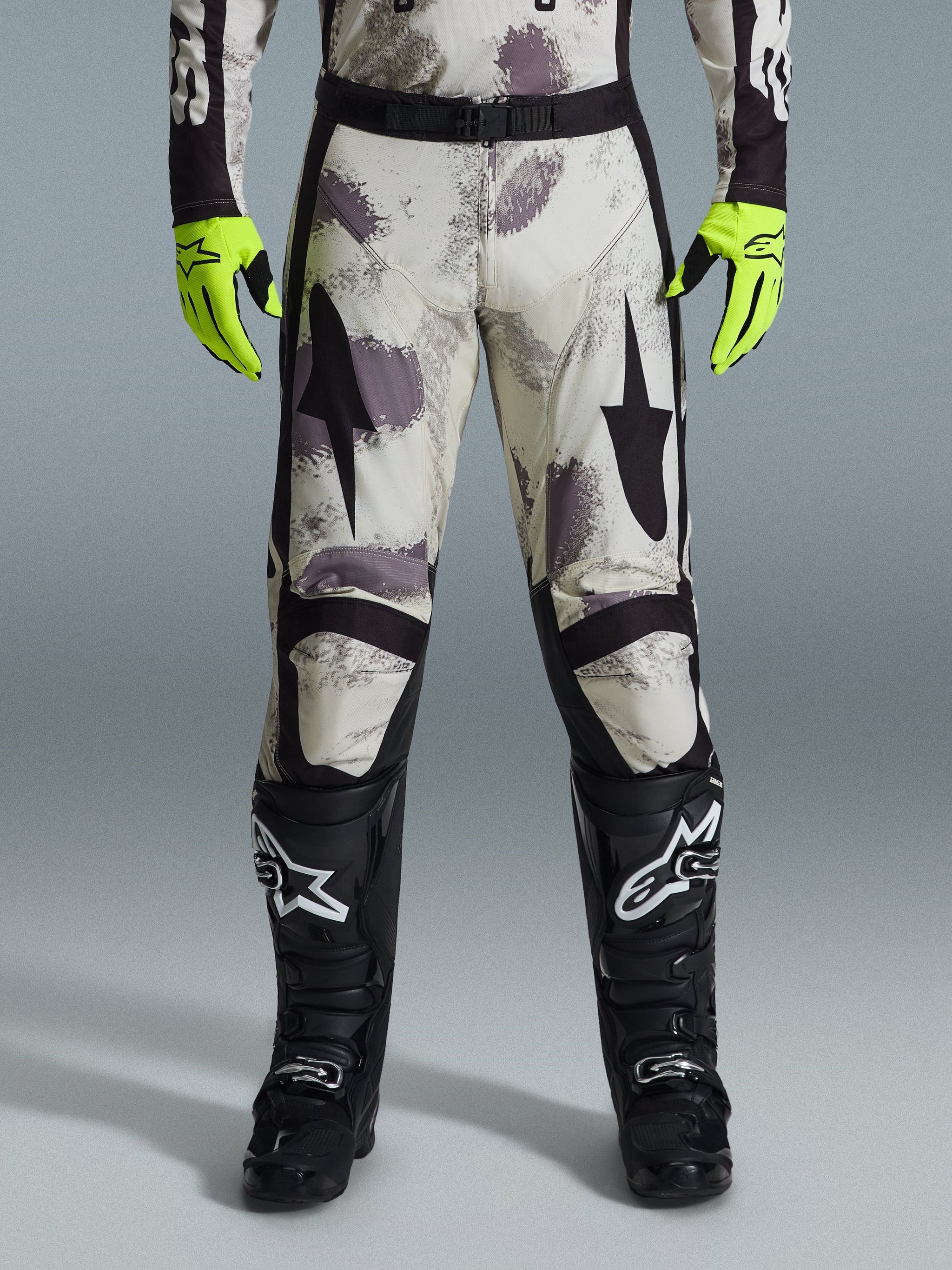 Spodnie Alpinestars 2026 Racer Lahnd, spodnie motocrossowe, wzór Sand Camo z szarymi i fioletowymi abstrakcyjnymi grafikami, widoczne z neonowożółtymi rękawicami i czarnymi butami, czarne logo gwiazdy na udach i czarny pas w talii, wytrzymały sprzęt do wyścigów off-road