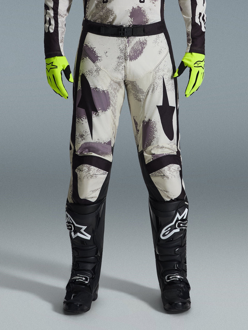 Spodnie Alpinestars 2026 Racer Lahnd, spodnie motocrossowe, wzór Sand Camo z szarymi i fioletowymi abstrakcyjnymi grafikami, widoczne z neonowożółtymi rękawicami i czarnymi butami, czarne logo gwiazdy na udach i czarny pas w talii, wytrzymały sprzęt do wyścigów off-road