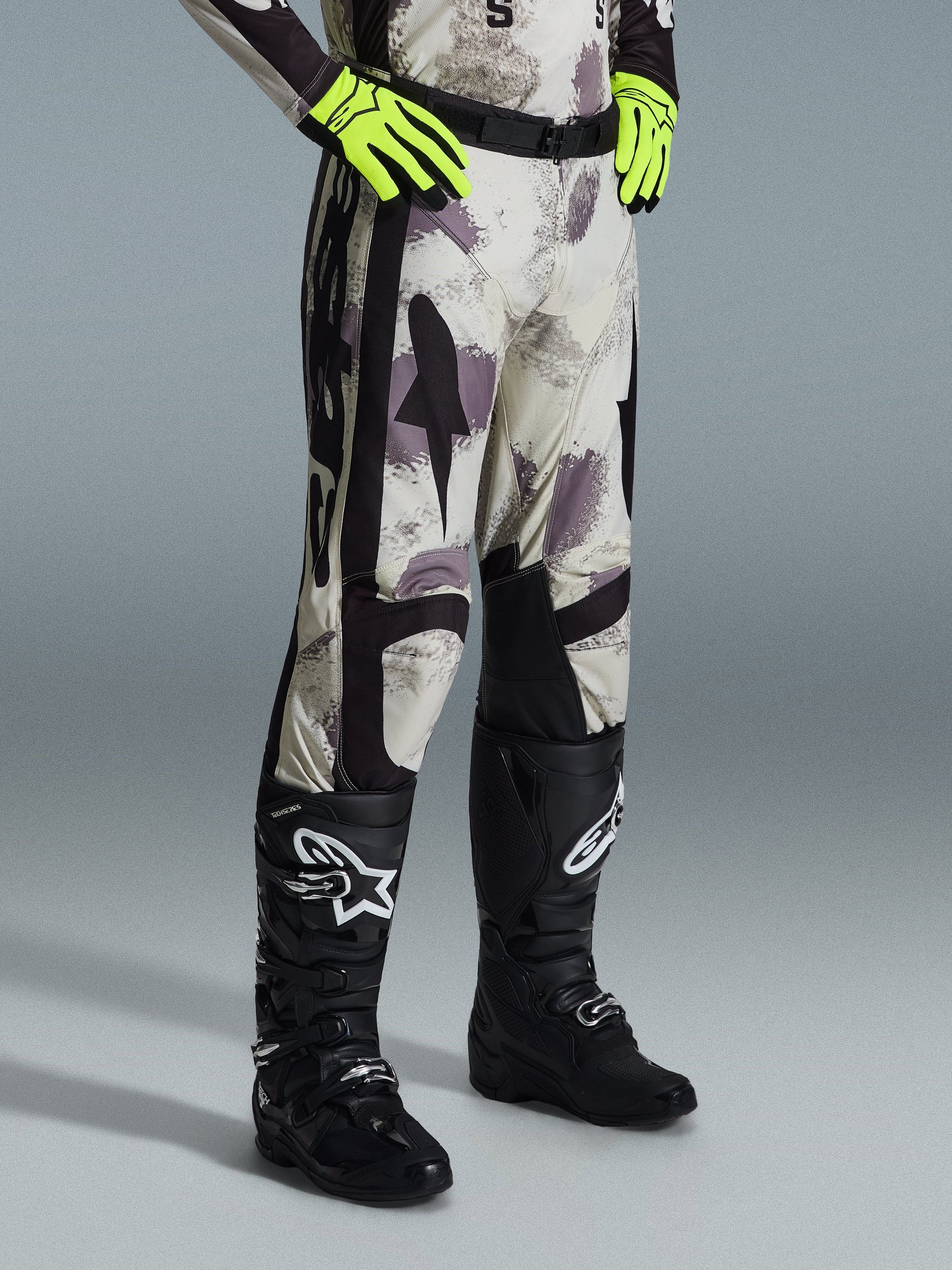 Alpinestars Racer Lahnd Pants 2026, spodnie motocrossowe, Sand Camo, zestawione z neonowożółtymi rękawicami i czarnymi butami motocrossowymi, z abstrakcyjnym wzorem kamuflażu i czarnymi panelami bocznymi, ergonomiczna konstrukcja off-road