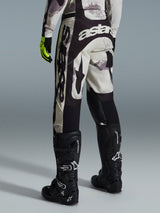 Alpinestars 2026 Racer Lahnd Pants, spodnie motocrossowe, kolor Sand Camo i szary, widok z tyłu z wyraźnym białym brandingiem na siedzeniu i nogawkach, w zestawieniu z czarnymi butami motocrossowymi i żółtymi rękawicami, profesjonalny sprzęt off-road