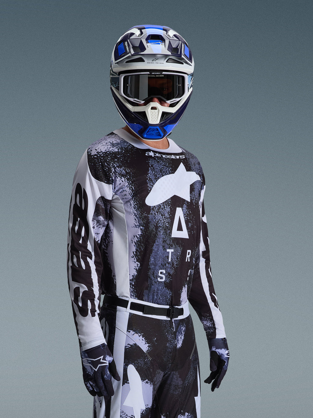 Motocyklista motocrossowy w zestawie Alpinestars 2026 Racer Lahnd, czarno-biała bluza z marmurkową niebiesko-szarą grafiką, pasujące spodnie i rękawice, niebiesko-biały kask z goglami, wyczynowy strój do wyścigów off-road