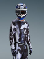 Motocyklista motocrossowy ubrany w koszulkę Alpinestars Racer Lahnd 2026 w kolorze Iron Camo, z czarno-szarym wzorem kamuflażu i dużym białym logo Alpinestars na piersi, w dopasowanym kasku i rękawicach do wyścigów terenowych.