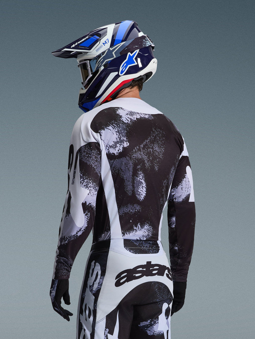 Koszulka Alpinestars 2026 Racer Lahnd w kolorze Iron Camo, widok z tyłu motocyklisty w czarno-biało-szarym zestawie odzieży z abstrakcyjnym wzorem, pasujące spodnie, czarne rękawice, niebieski kask wyścigowy i gogle.