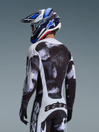 Koszulka Alpinestars 2026 Racer Lahnd w kolorze Iron Camo, widok z tyłu motocyklisty w czarno-biało-szarym zestawie odzieży z abstrakcyjnym wzorem, pasujące spodnie, czarne rękawice, niebieski kask wyścigowy i gogle.
