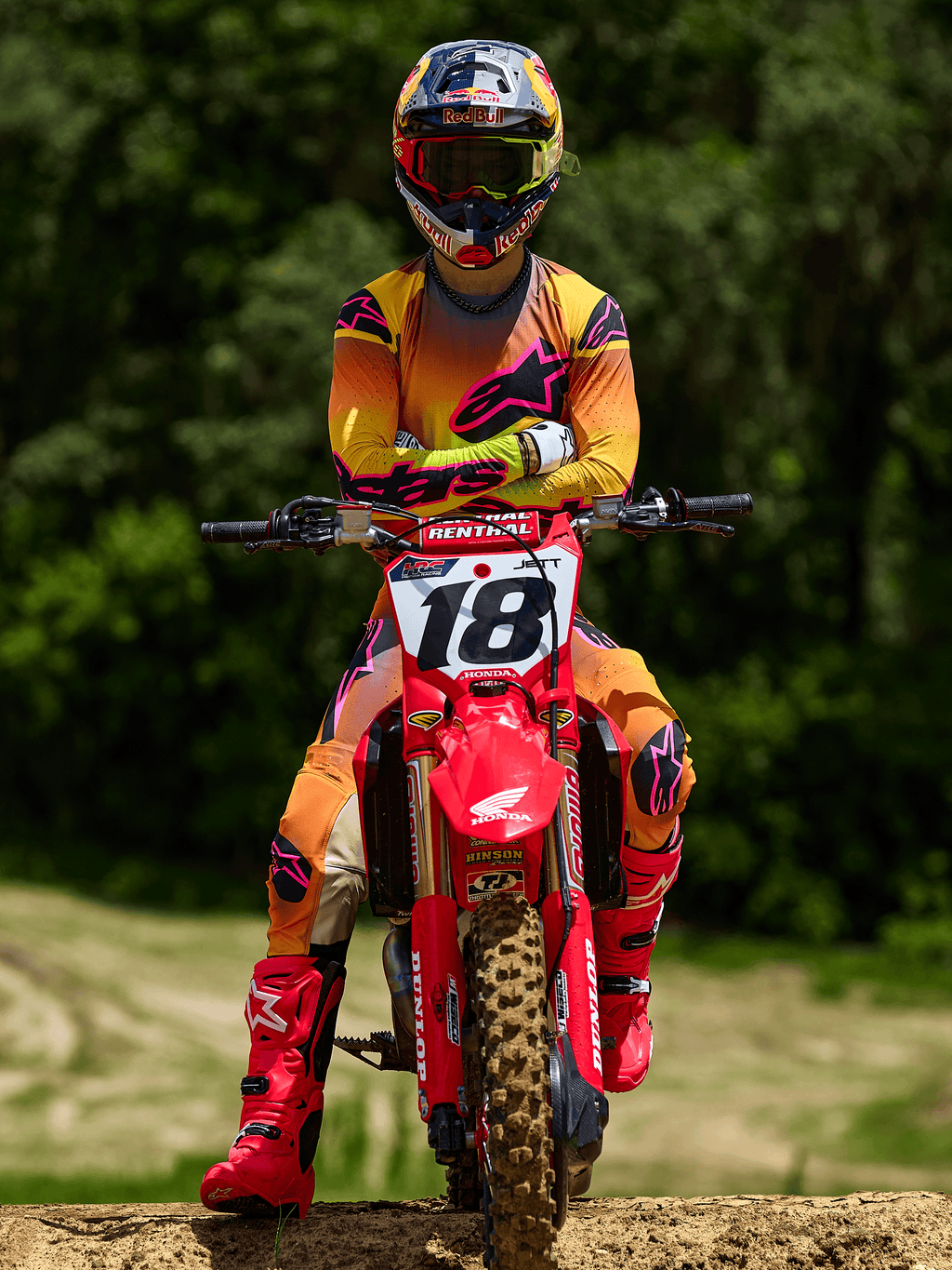 Motocyklista motocrossowy siedzący na czerwonym motocyklu crossowym Honda z numerem startowym 18, ubrany w koszulkę Alpinestars 2025 Supertech Lipan i pasujące spodnie w kolorze żółto-różowym, jaskraworóżowe buty Alpinestars, kask Red Bull, profesjonalny strój wyścigowy do jazdy terenowej.