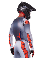 Motocyklista motocrossowy widoczny od tyłu w koszulce Alpinestars 2025 Supertech Lipan w kolorze Gray Orange Fluo, z wyraźnymi pomarańczowymi logotypami na rękawach i plecach, w zestawie z dopasowanymi spodniami i czarnym kaskiem, profesjonalny strój wyścigowy do jazdy w terenie.
