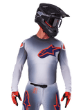 Alpinestars Supertech Lipan Jersey 2025, koszulka motocrossowa, szara z pomarańczowymi i granatowymi akcentami, techniczna koszulka z długim rękawem i perforowanymi detalami wentylacyjnymi, noszona z czarnym kaskiem i czerwonymi rękawicami, nowoczesny design wyścigowy