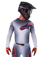 Alpinestars Supertech Lipan Jersey 2025, koszulka motocrossowa, szara z pomarańczowymi i granatowymi akcentami, techniczna koszulka z długim rękawem i perforowanymi detalami wentylacyjnymi, noszona z czarnym kaskiem i czerwonymi rękawicami, nowoczesny design wyścigowy