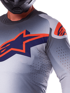 Alpinestars 2025 Supertech Lipan Jersey, koszulka motocrossowa, szaro-pomarańczowa fluo, z dużym granatowo-pomarańczowym logo gwiazdy na piersi, laserowo wycinane perforacje zapewniające przepływ powietrza, lekka konstrukcja z siatki, noszona z czarnym kaskiem