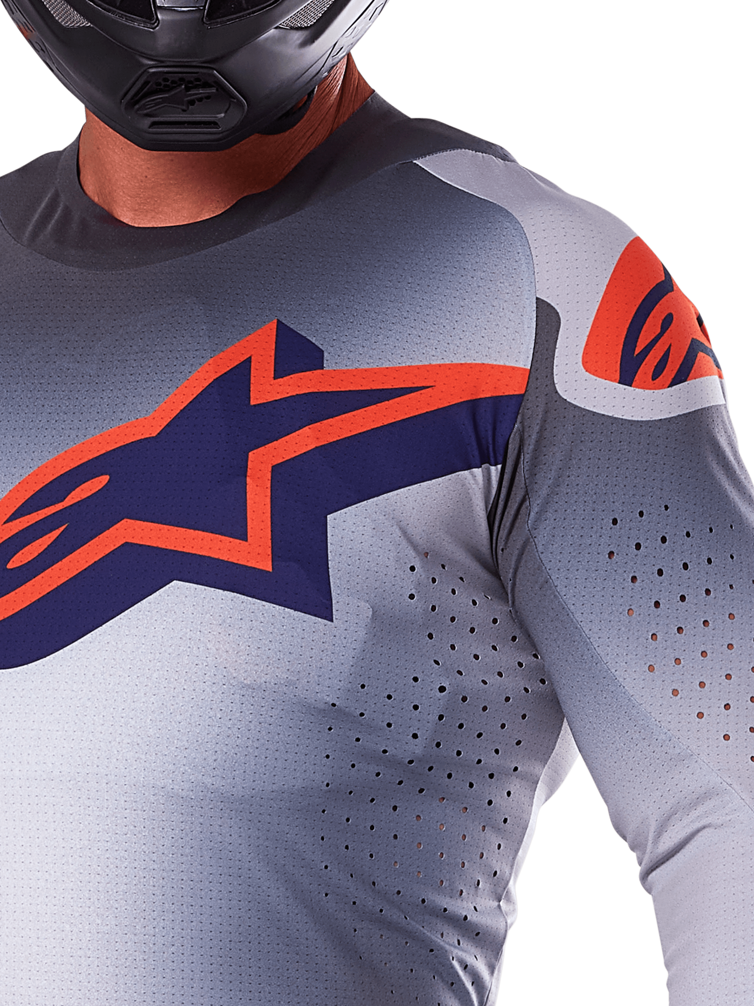 Zbliżenie na zawodnika w koszulce Alpinestars 2025 Supertech Lipan, koszulka motocrossowa, szara i pomarańczowa fluo, z dużym pomarańczowym logo Alpinestars na klatce piersiowej, laserowo wycinanymi otworami wentylacyjnymi i czarnym kaskiem, profesjonalny sprzęt wyścigowy.