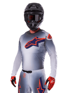 Motocyklista motocrossowy w zestawie Alpinestars 2025 Supertech Lipan, szara koszulka i spodnie z granatowymi i pomarańczowymi akcentami, matowy czarny kask z goglami oraz czerwono-czarne rękawice, sportowa odzież wyczynowa do wyścigów terenowych