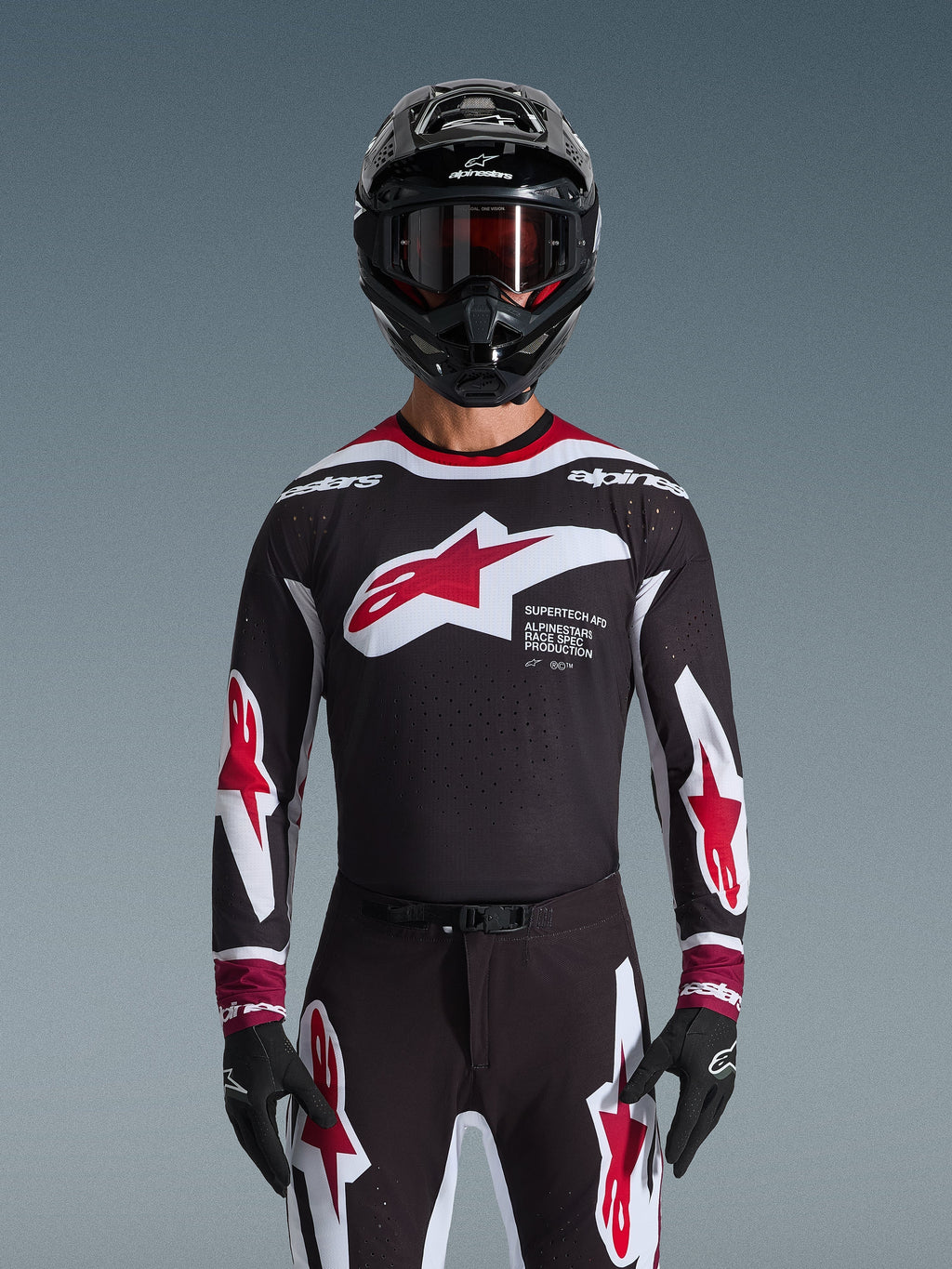 Alpinestars 2026 Supertech AFD Vista Jersey, koszulka motocrossowa w kolorach czarnym, czerwonym i białym, widoczna na motocykliście w czarnym kasku, goglach i rękawicach, z dużym logo na piersi i brandingiem „SUPERTECH AFD”, profesjonalna odzież wyścigowa do jazdy terenowej