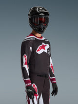 Motocyklista motocrossowy ubrany w koszulkę Alpinestars 2026 Supertech AFD Vista w kolorze czarno-czerwono-białym, z dużym czerwonym logo gwiazdy na piersi i laserowo wycinanymi perforacjami, w połączeniu z czarnym kaskiem i dopasowanymi spodniami wyścigowymi.