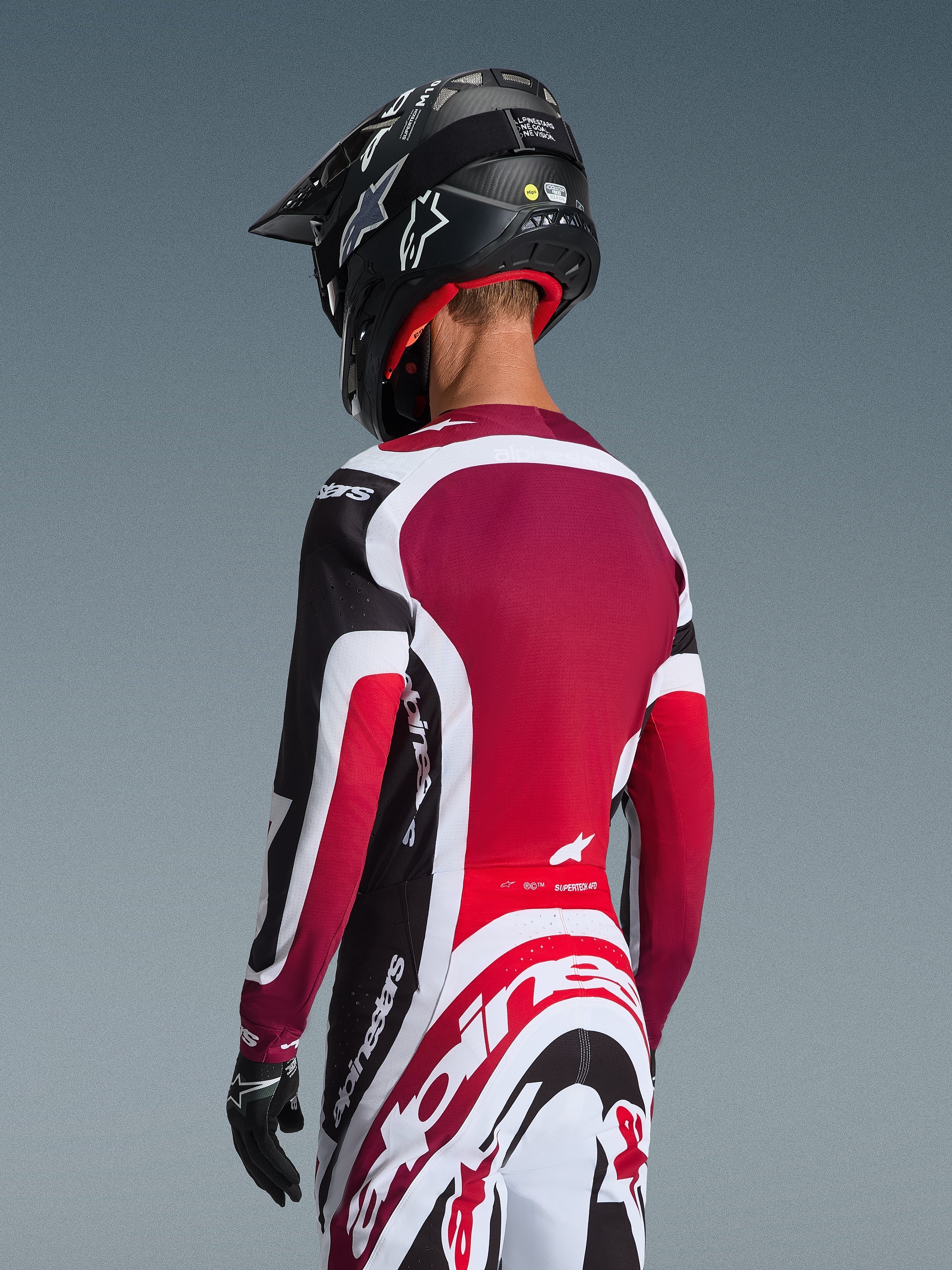 Widok z tyłu motocyklisty motocrossowego ubranego w koszulkę 2026 Supertech AFD Vista w kolorze czarno-czerwono-białym, zestawioną z dopasowanymi spodniami i czarnym kaskiem, z wyraźnym brandingiem Alpinestars, czerwonymi panelami z siatki i sportowym krojem wyścigowym.
