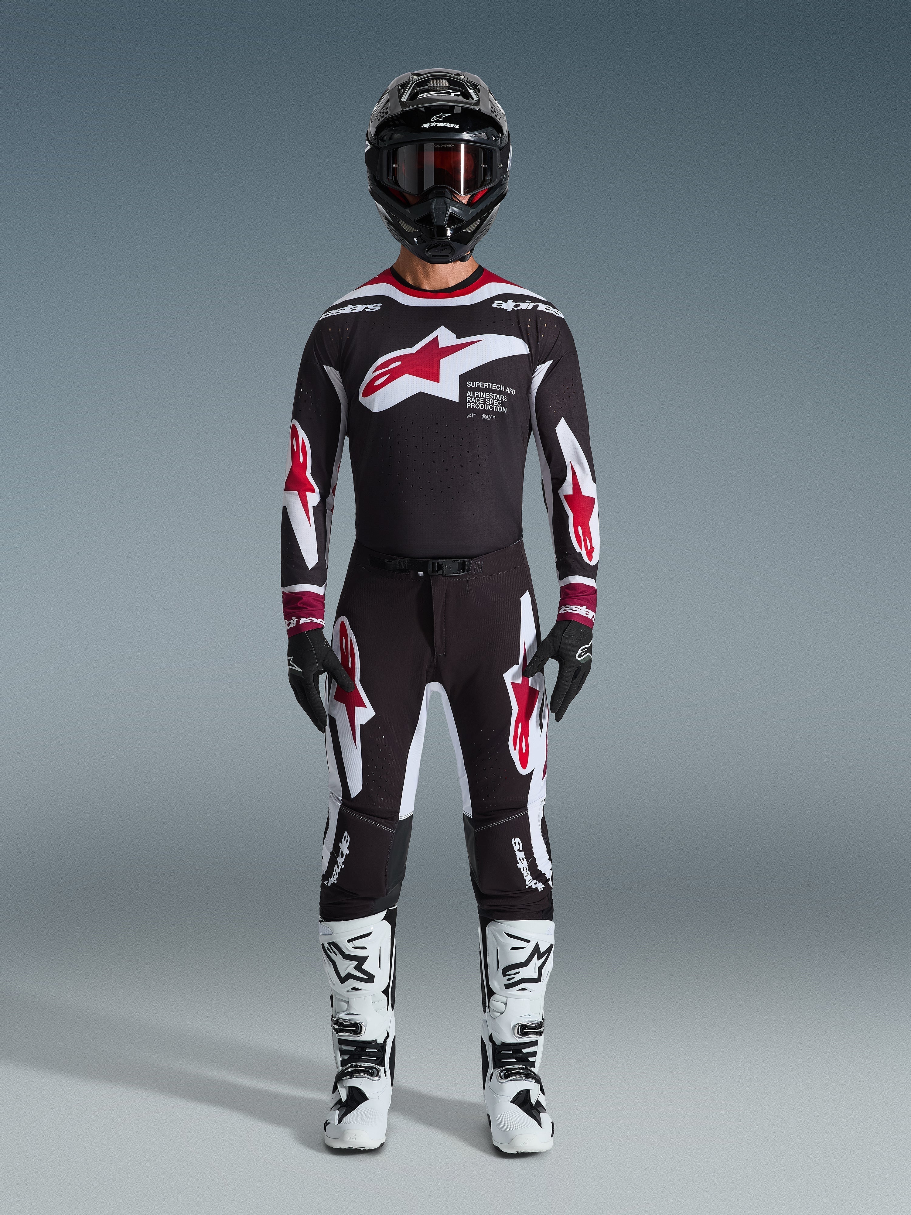 Zawodnik motocrossowy w koszulce Alpinestars 2026 Supertech AFD Vista, czarno-czerwono-biały strój wyścigowy, czarne spodnie z czerwonymi logotypami gwiazdy, biało-czarne buty, czarny kask z goglami, profesjonalny zestaw do jazdy w terenie