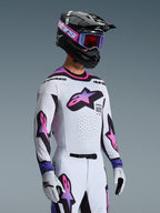 Kierowca motocrossowy w zestawie Alpinestars 2026 Supertech AFD Vista, biała bluza i spodnie z różowo-fioletowymi gradientowymi logotypami gwiazdy, czarny kask z fioletowymi akcentami, fioletowe gogle z czerwonymi szybkami i dopasowane białe rękawice, profesjonalny strój wyścigowy off-road.